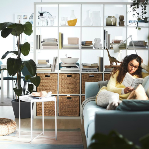 IKEA’s KALLAX white shelf unit and a large potted palm plant are used as room dividers between the dining area and the living room where a woman sits on a couch reading a book