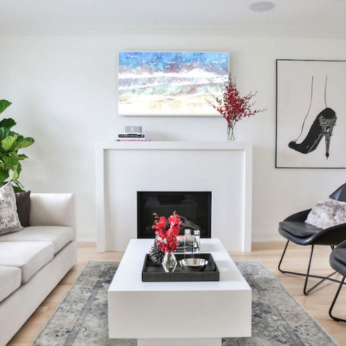 A chic white living room with a cozy gas fireplace surrounded by a custom-built mantle made of Laminam porcelain, an art TV and a large framed artwork on the wall, two black leather occasional chairs, a large white rectangular coffee table on a patterned grey rug, a large potted fig plant, and a white couch with grey throw pillows