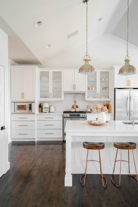 An all-white-everything kitchen featuring high ceiling, white walls, white cabinetry with sleek brass hardware, two sets of frosted glass cabinets, industrial silver pendant lamps, dark wooden floors and matching dark wooden barstools.