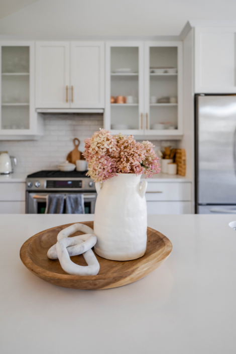 Atop the white kitchen island, a decorative wooden tray holds a white decorative chain-link sculptural piece and a white ceramic vase housing fresh pink flowers.