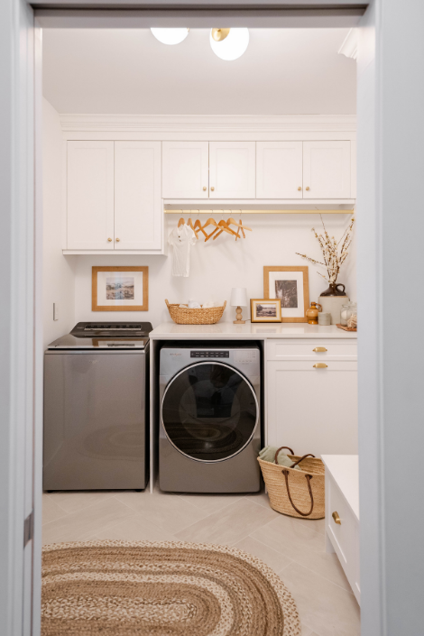 A laundry room with white cabinets, a wood railing holding wooden hangers, a grey washer and dryer set and plenty of wood and wicker touches.