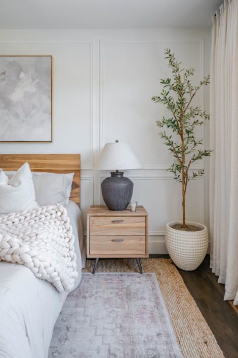 A tall faux olive tree in an off-white ceramic planter stands in a corner of the bedroom, by the bedside table.