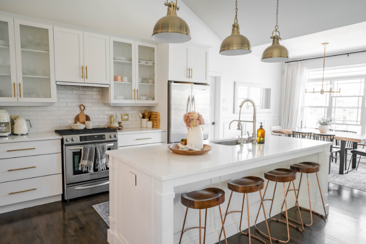 A large white kitchen island sits in the middle of the kitchen with a row of dark wooden barstools tucked beneath it. Large silver industrial-style pendant lamps hang overhead.