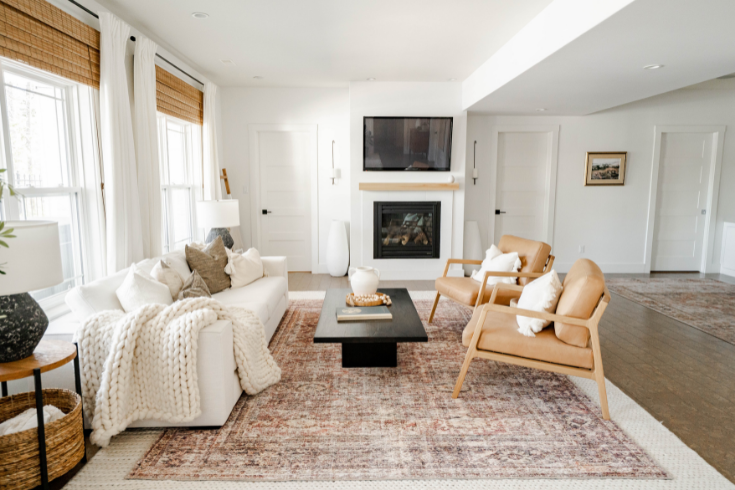 A TV room featuring white walls, large windows with white curtains and wicker blinds, a white sofa, a pair of matching wooden armchairs with peachy-brown upholstery and a TV mounted above a fireplace.