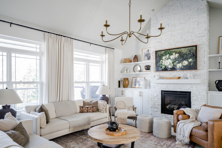 Large windows with long white drapes line one wall of the white living room. The room is furnished with off-white matching sofas, an off-white armchair, a tan leather armchair and a round wood and iron coffee table.