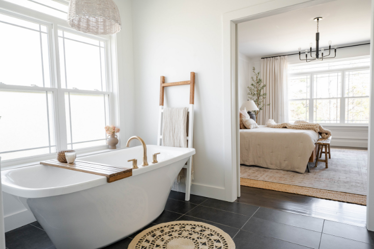 A wide doorway from the principle bedroom looks into a spacious bathroom with white walls, black tile floors and a large white standup tub.