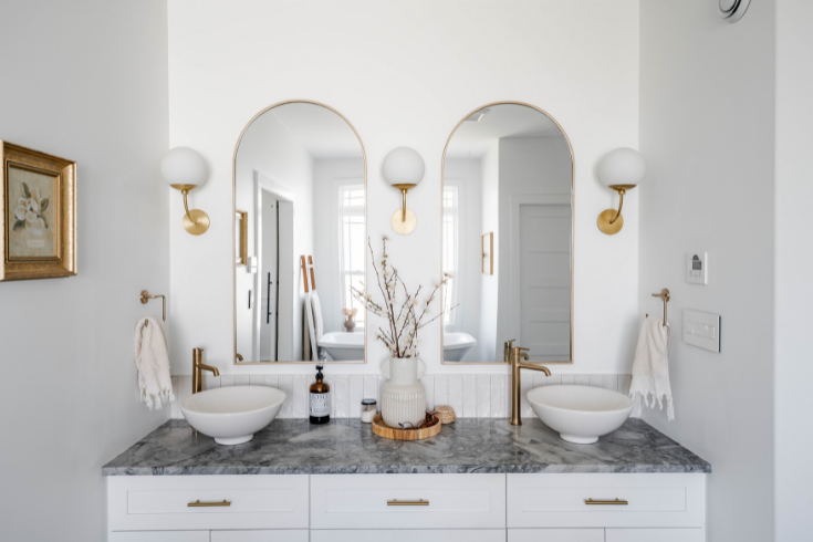 Arched mirrors are mounted atop a white vanity with a dark counter. Brass wall sconces are mounted on either side of and in between the double mirrors.