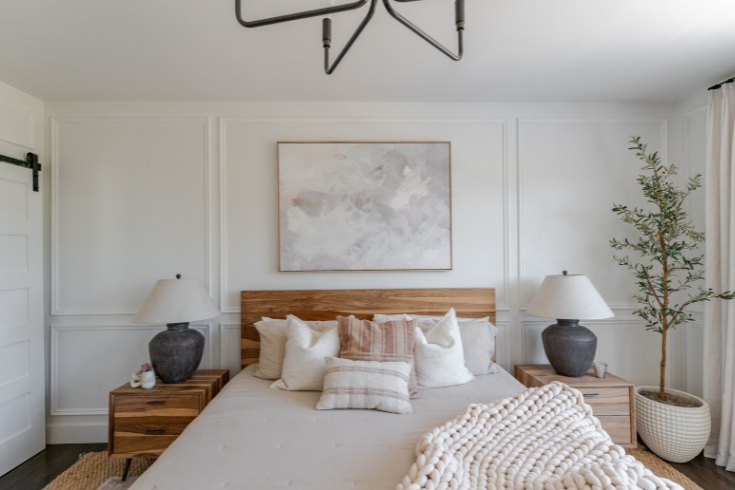 The primary suite, featuring white walls, a large wooden bed with matching wooden bedside tables, a large piece of art above the bed's headboard and plush bedding in a palette of white and neutrals.