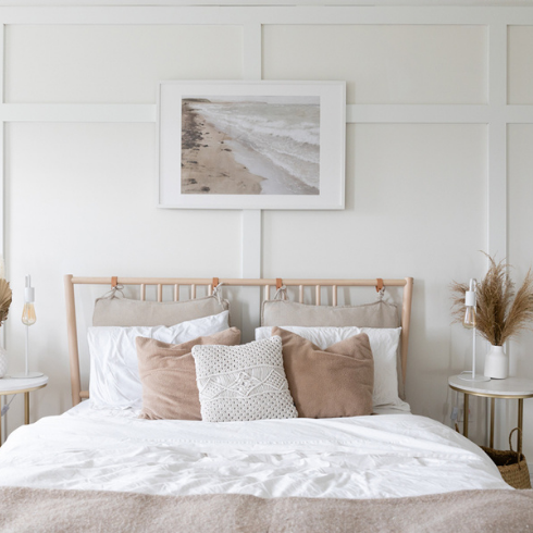 Lauren Sheriff's home tour showing her principle bedroom: Clean white walls with picture frame molding, a wooden bed outfitted in neutral coloured bedding and a serene art piece in neutral colours hanging over the bed's headboard.