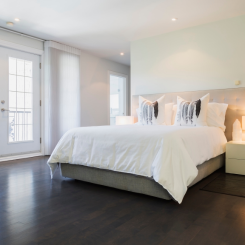 A large, clean bedroom with empty white walls, a bed outfitted in a cushy white duvet and dark wooden floors with no carpeting, rugs or clutter in the way.