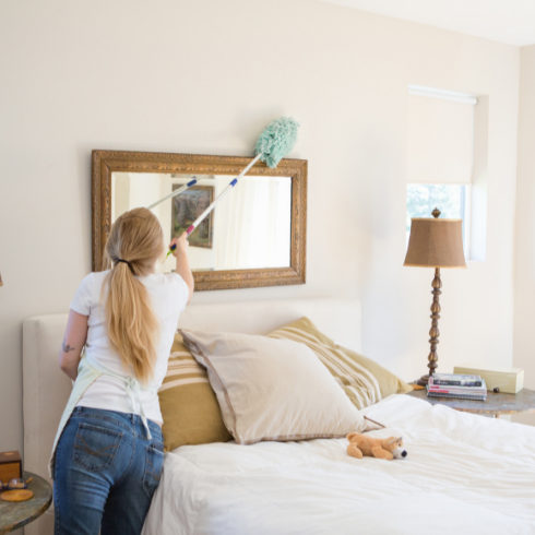Woman in white t shirt an d jeans using a long-handled duster to dust around the gilded frame of a mirror hanging above her bed.