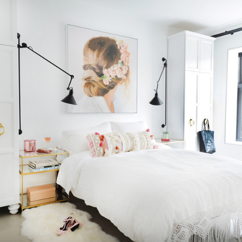 A clean aesthetic bedroom featuring all white walls, white cabinetry, white bedding and a white shag bedside rug. Above the bed is a large pink and white piece of art featuring a woman wearing a flower crown. The bed is decorated with pink and white cushions. Black wall-mounted bedside lamps flank the bed.