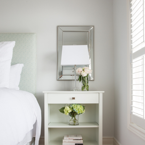 White bedside table with square drawer pull, two floral arrangements, three books on lower shelf