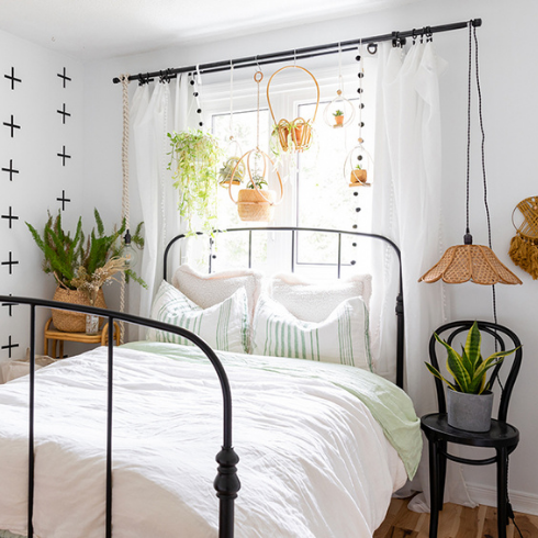 A bedroom with bright white walls, an accent wall patterned with black crosses, a black iron bed with crisp white bedding and lots of indoor plants. Six hanging baskets of plants hang from the curtain rod at the window behind the headboard while two larger potted plants flank the bed.