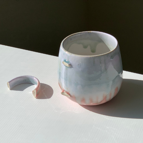A blue, white and pink ceramic mug with a broken handle. Both mug and handle sit atop a white counter.