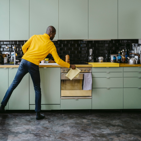 A man in a yellow sweater and jeans cleaning his sage-green kitchen cabinets with a rag.