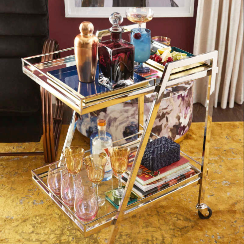 A chrome bar cart with glass shelves filled with decanters, glassware and a martini shaker
