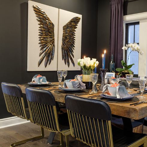 A dining room with black walls, a large wooden dining table with six chairs, and artwork of two large wings.