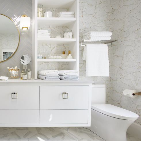 A white floating vanity, wall shelves, round gold mirror, two gold vanity lights, and marble walls in a chic bathroom designed by Michael Holmes and Jo Alcorn featured on HGTV’s Home to Win: For the Holidays