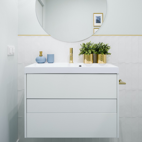 White bathroom with a simple white floating and a touchless gold faucet, with a round mirror above it on the wall