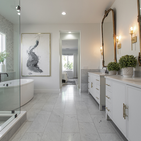 Large white bathroom with rectangular marble floor tiles, a white free standing tub, two white vanities, a large gold framed print on the wall looking into a bedroom as featured on HGTV’s Rock The Block