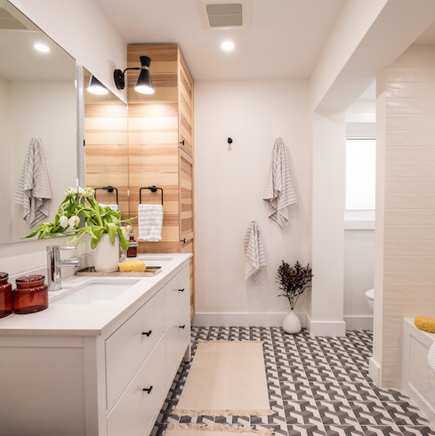 Jonathan and Drew Scott designed this spa-like bathroom for HGTV’s Property Brothers: Forever Home featuring tile flooring in a fun, modern pattern, wooden built-in closets, a large white double sink vanity and a black sconce light