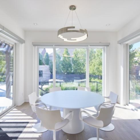 A modern dining room with a circular white dining table and white chairs, surrounded by floor-to-ceiling windows.
