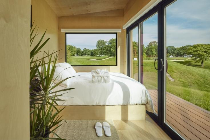 The interior of a one-bedroom pop-up hotel with a white bed looking onto a golf course