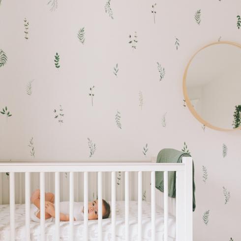 A baby in a nursery with a white crib, white wall with green leaf decals