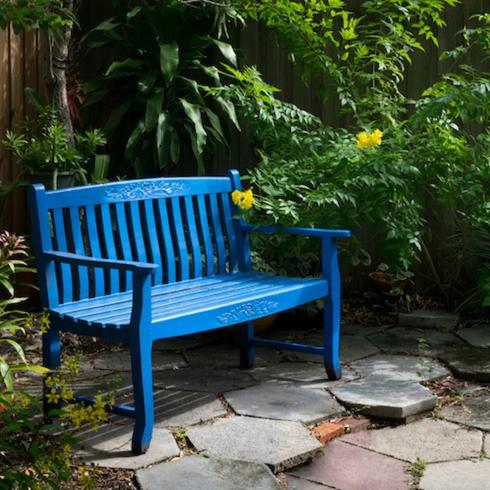 Blue bench sit on a hexagon tiled ground in a lush garden with palm trees and yellow flowers in a memorial garden
