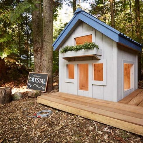An adorable dog-sized mini cottage in the woods with a sign that says “Welcome Crystal” out front