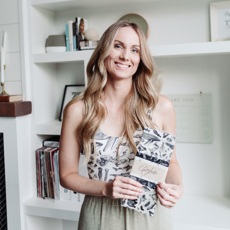 Amy Hall founder of Goldilocks Goods stands in front of a white bookcase holding a set of Goldilocks Goods beeswax wraps