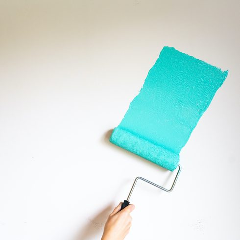 Paint roller applying teal coloured paint to white wall as part of a painting starter kit of essential tools