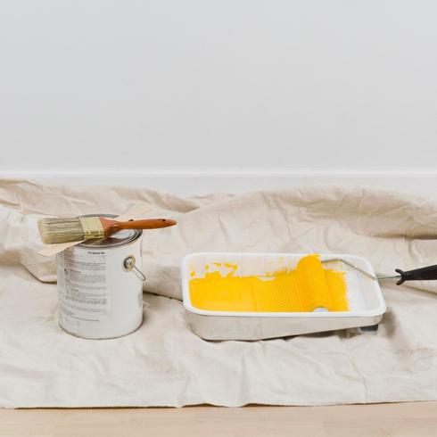 Part of painting essential tools a paint can, paint brush and roller and tray with yellow paint sit on a cotton drop cloth on a wood floor against a white wall