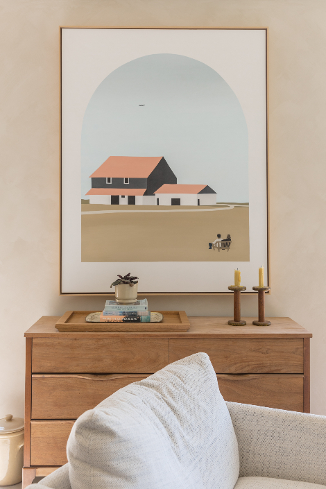 A framed art print hanging above a wooden sideboard with a white armchair in the foreground