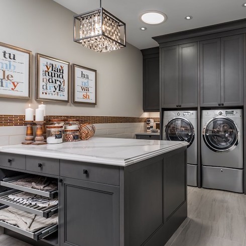 Spacious and fully functional laundry room with grey cabinets, a centre island with built-in drying racks, a crystal chandelier and large grey tile floors as part of a home reno trends story for HGTV Canada
