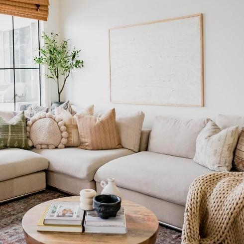 Large taupe corner sectional couch with piles of organic looking pillows in a white boho chic living room with large windows and a chunky knitted blanket as part of a home reno trends story for HGTV Canada