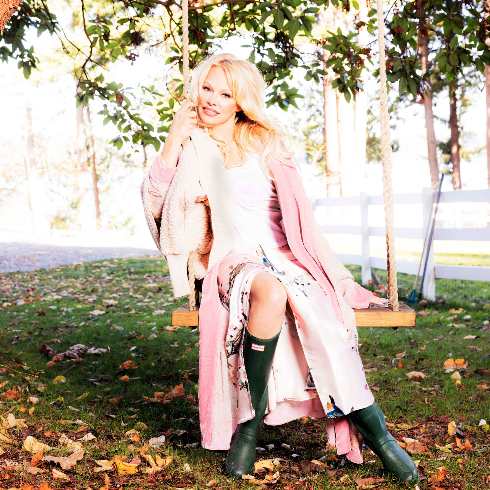 Pamela Anderson on a wooden swing in white dress and pink coat