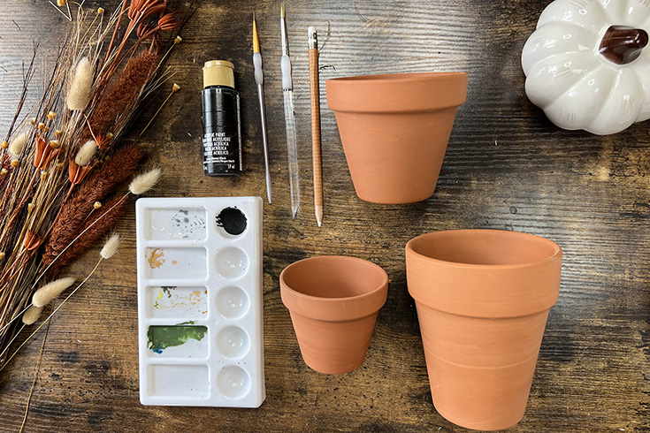 An overhead shot of required materials – terracotta pots, black acrylic paint, two paint brushes, a chalk pencil, and a paint palette. 
