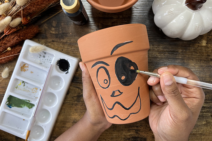 Person's hands painting a larger terracotta pot with a goofy jack-o-lantern face