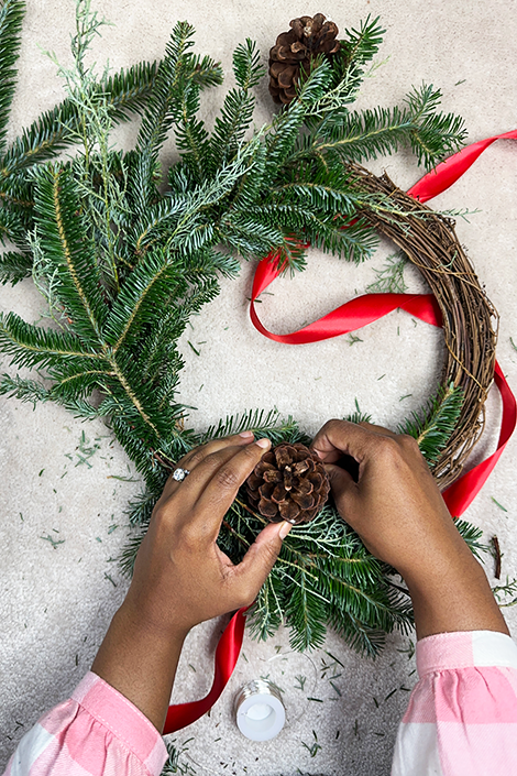 Hands attaching a pinecone to a nearly-finished green wreath - wreath tutorial
