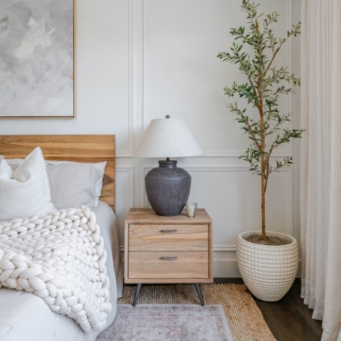 One side of a bed with chunky knit blanket, wooden side table and large plant in the corner