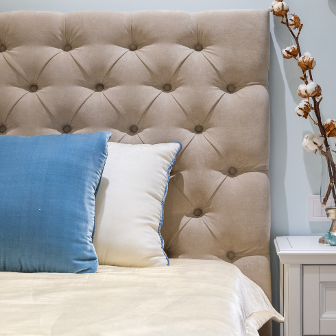 A taupe upholstered headboard with neutral bedding, a blue accent pillow and a white accent pillow