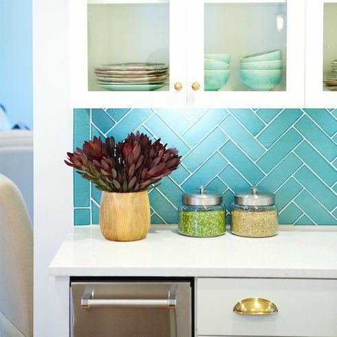 Turquoise glass backsplash tiles in a herringbone pattern adds a sense of beachy calm to this bright white kitchen. Coordinating dishware in the glass-fronted cabinets above help reinforce the coastal aesthetic.