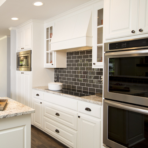 White modern kitchen with two wall ovens and stovetop with a high shine black tile backsplash