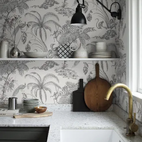 Toile de Jouy wallpaper Moa in Sandstone by Sandberg Wallpaper features monkeys swinging between the palm trees are accompanied by turtles, peacocks, and beautiful flowers on the wall of a simple chic kitchen with an open shelf and a brass tap