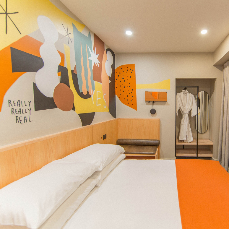 A hotel room with a bright mural and orange and white sheets