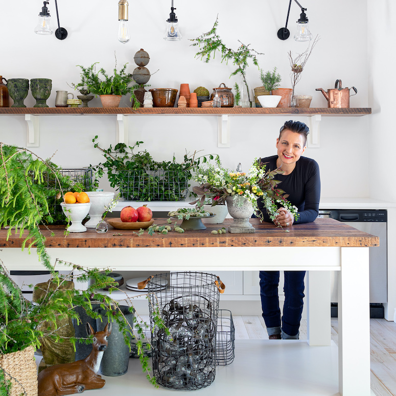 Designer Cynthia Zamaria stands behind a large butcher block counter top island surrounded by flowers, fruits and antiques
