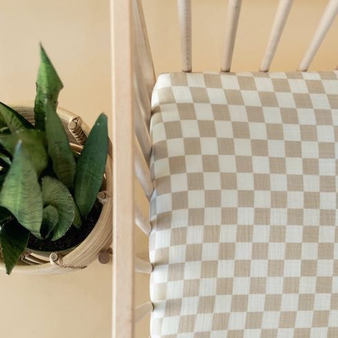 Checkerboard Crib Sheet by Brixton Phoenix in a pine wood crib next to a potted plant