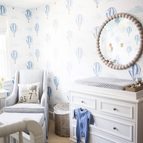 A nursery room with a mirror, drawers and white and blue wallpaper with hot air balloon design